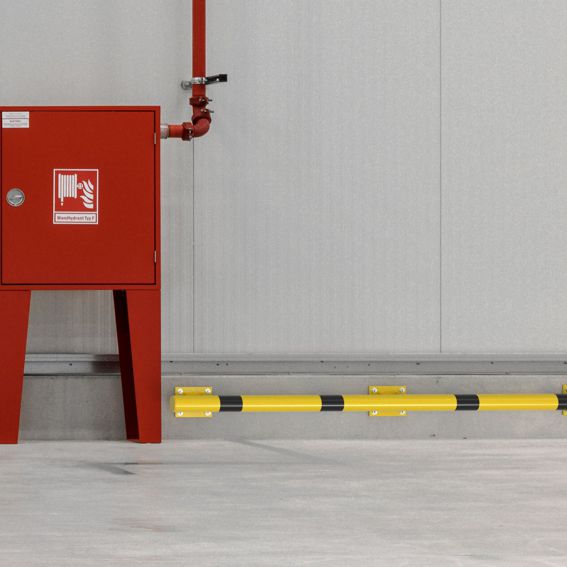 Gelb-schwarzer Rammschutzbalken an einer Wand schützt einen Feuerlöschkasten in einer Industriehalle.